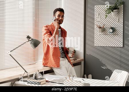 Porträt eines selbstbewussten, hübschen jungen schwarzen Mannes in stilvollem Outfit, der mit Kaffee zum Mitnehmen am Fenster steht und im Büro telefoniert Stockfoto