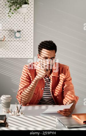 Seriöse junge Mixed-Race-Expertin für Brillen, die am Schreibtisch sitzt und Bleistift hält, während sie mit Papieren arbeitet Stockfoto