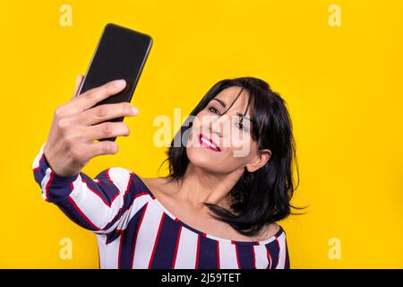Die schöne Frau trägt ein gestreiftes Hemd. Die junge Brünette nimmt ein Selfie mit ihrem Mobiltelefon auf gelbem Hintergrund auf. Stockfoto