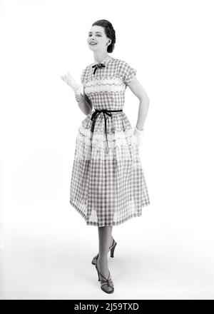 1950S BRÜNETTE MODELL TRÄGT KARIERTES KURZARM-KLEID MIT SPITZEN-BANNER AUF ROCK UND BÜSTE BAND KRAWATTE AN TAILLE UND HALS - F3873 CRR001 HARS BÜSTE UND KARIERTE BERUFE POSIEREN STILVOLLE MODELLIERUNG MODE MODELLIERUNG POSE TAILLE JUNGE ERWACHSENE FRAU BANNER SCHWARZ UND WEISS ALTMODISCH Stockfoto