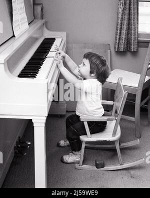 1960S MÄDCHEN KLEINKIND SITZT IN CHILD’S SCHAUKELSTUHL AN EINEM KLAVIER IHRE FINGER NUR ERREICHEN SPITZEN DER TASTEN SUCHEN BEI NOTEN - J11787 HAR001 HARS COPY SPACE FULL-LENGTH INSPIRATION JUST B&W BRUNETTE TRÄUMT HIGH-ANGLE GELEGENHEIT MUSIKINSTRUMENT NEUGIERIGE CHILD'S TIPPS WACHSTUM JUGENDLICHE TALENT BABY MÄDCHEN SCHWARZ UND WEISS KAUKASISCHEN ETHNIZITÄT HAR001 ALTMODISCH Stockfoto
