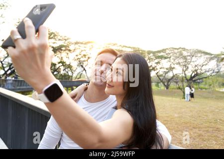 Asiatische Frau, die während des Sonnenuntergangs mit seinem Mann in einem Park ein Sefie gemacht hat Stockfoto