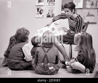 1970S LEHRERIN LIEST EINER GRUPPE VON JUNGEN UND MÄDCHEN AUS DEM KINDERGARTEN EIN BUCH VOR, DIE AUF DEM BODEN SITZEN HER - J13443 HAR001 HARS ALTE MODE SCHWESTER 1 JUGENDLICHE GRUNDSCHULLEHRER KOMMUNIKATION JUNGE ERWACHSENE FREUDE LIFESTYLE FRAUEN BRÜDER STUDIO SCHUSS 6 KOPIE RAUM HALBE LÄNGE DAMEN PERSONEN INSPIRATION MÄNNER SECHS GESCHWISTER VERTRAUEN SCHWESTERN B&W ZIELE SCHULEN GRAD GLÜCK KINDERGARTEN VORSCHULE ENTDECKUNG UND WAHL AUFREGUNG LEHRER FÜHRUNG LOW-ANGLE GELEGENHEIT BERUFE PRIMÄRE GESCHWISTER VERBINDUNG KONZEPTIONELLE STILLLEBEN ERZIEHER VORSCHULE TAGESBETREUUNG BILDBAND ERZIEHEN Stockfoto