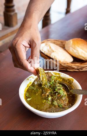 Ein Stück Goan-Brot wird von Menschenhand in ein authentisches Goan-Gericht namens Chicken Cafreal getaucht. Stockfoto