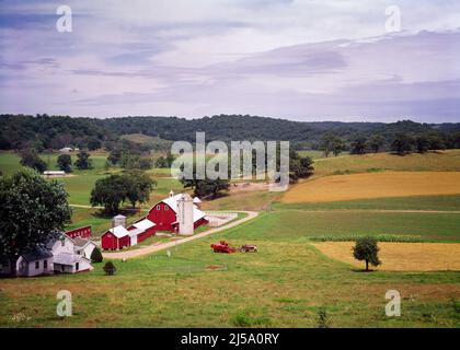 1970S LANDSCHAFTLICH SCHÖNE AUSSICHT AUF FAMILIENFARM MIT BAUERNHAUS ROTEN SCHEUNEN SILO UND TRAKTOR AUF DEM FELD SANFT ROLLENDE HÜGEL WISCONSIN USA - KF7816 VGE001 HARS IMMOBILIEN STRUKTUREN BAUZEIT SILO SCHEUNEN ALTMODISCH Stockfoto