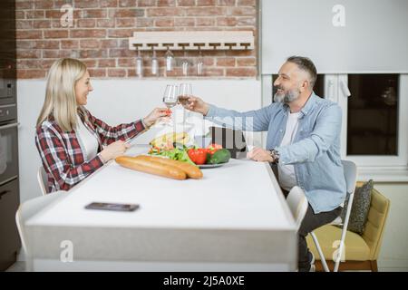 Glückliches reifes Ehepaar, das am Küchentisch sitzt und mit Gläsern Wasser toaste. Frisches Bio-Gemüse für das Gericht vorbereitet. Gesunde Ernährung und Lebensstil Konzept. Stockfoto