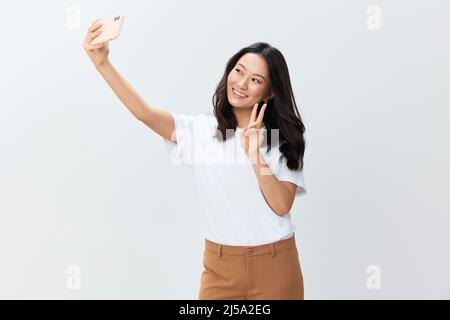 Lasst mich ein Selfie machen. Attraktive fröhliche asiatische junge Frau in weißem Grund T-Shirt tun Schuss mit Telefon posiert isoliert auf weißem Studio Hintergrund Stockfoto