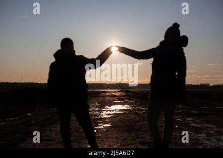 Mädchen zeigen ihre Herzen vor dem Hintergrund der Sonne. Zwei Frauen halten ihre Hände zusammen. Symbol der Freundschaft. Freunde am Himmel. Silhouetten von Menschen. Stockfoto