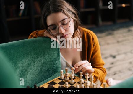 Nahaufnahme einer nachdenklichen, niedlichen jungen Frau mit eleganten Brillen, die Schach in Bewegung bringen und ein Ritterstück auf dem Holzboden sitzen lässt. Stockfoto