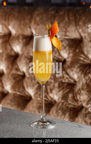 Glas eines köstlichen gelben Cocktails French 75 steht auf der Theke auf dem verschwommenen dunklen und gelben Hintergrund. Stockfoto