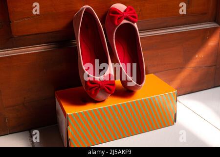 Hübsche flache Schuhe für Mädchen, rosa, pinkes Band, an Holztür gelehnt, gelb gestreifte Boxsohle Stockfoto