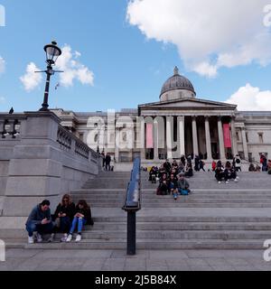 Touristen sitzen entspannt und checken auf der Treppe am Trafaglar Square vor der National Gallery, London. Stockfoto