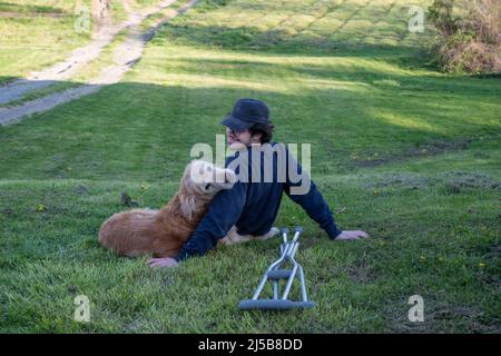 Hund kuschelt mit seinem Besitzer auf einem ländlichen grasbewachsenen Hügel mit einem Paar Krücken auf dem Boden im Vordergrund. Der Hund blickt im Freien zurück zur Kamera Stockfoto