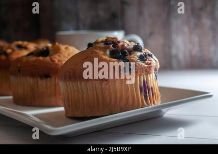 Heidelbeer-Muffins auf einem weißen quadratischen Teller mit einer Tasse Heißgetränk Kaffee Kakao Schokolade und einer Holzwand im Hintergrund, Stockfoto