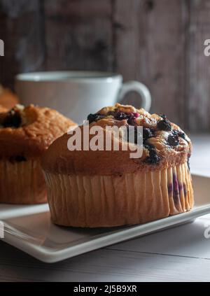 Heidelbeer-Muffins auf einem weißen quadratischen Teller mit einer Tasse Heißgetränk Kaffee Kakao Schokolade und einer Holzwand im Hintergrund, Stockfoto