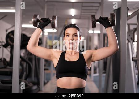 Portrait kaukasische Frau im Fitnessstudio trainiert ihre Schultern mit Kurzhanteln in ihren Händen, macht Overhead-Presse, sitzt auf einer Bank Stockfoto