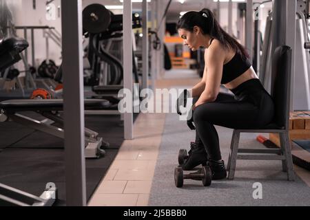 Die athletische Frau sitzt und ruht sich nach dem Training oder dem Training im Fitnessraum aus. Entspannungskonzept. Das Thema Krafttraining und Bodybuilding. Stockfoto