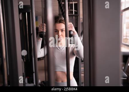 Junge Frau trainiert mit Gewichten im Fitnessstudio. Übungen am Simulator mit Gewichten. Das Mädchen führt die Pull-Down-Übung durch. Stockfoto