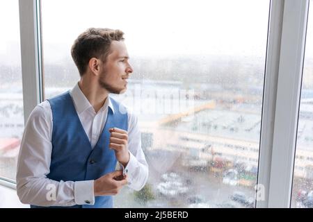 Portrait ein arroganter, scharf gekleideter Mann in blauem Anzug korrigiert Knopf auf Manschettenknöpfen sein Hemd, bereitet sich auf das Date Meeting vor, schaut lächelnd zum Fenster hinaus und erwartet eine erfolgreiche Transaktion Stockfoto