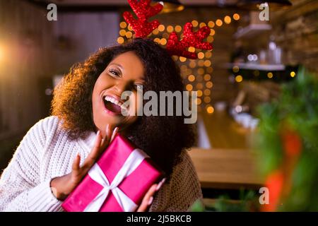 brasilien Brünette Haare Frau halten rote Geschenkbox in der Nähe von Tannenbaum in ihrer Wohnung ist sie tragen weißen Pullover und rote Hirsch Hörner Stockfoto