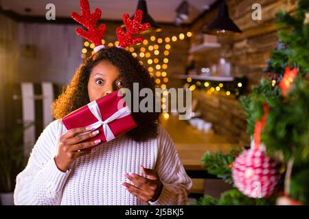 brasilien Brünette Haare Frau halten rote Geschenkbox in der Nähe von Tannenbaum in ihrer Wohnung ist sie tragen weißen Pullover und rote Hirsch Hörner Stockfoto