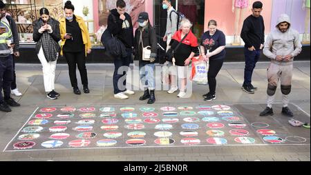 Ein Straßenpflaster-Künstler, der einen grauen Trainingsanzug trägt, zeichnet Bilder von Flaggen oder Flaggen vieler Länder der Welt auf einem öffentlichen Bürgersteig im Zentrum von Manchester, England, Großbritannien und den Britischen Inseln. Passanten hinterlassen Münzen auf den Flaggen, die ihnen gefallen. Stockfoto