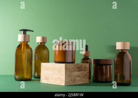 Flaschen für Kosmetik und Creme auf einem Sockel, grüner Hintergrund. Braune Glasgläser mit Holzdeckeln. Eco-Konzept. Behälter für Shampoo, Öl. Flasche mit Spender und Pipette. Stockfoto