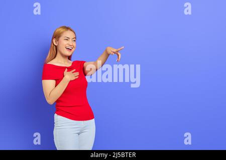 Porträt einer fröhlichen asiatischen Frau, die lacht, während sie isoliert auf violettem Hintergrund zur Seite zeigt Stockfoto