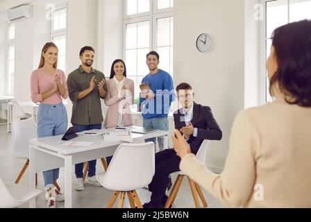 Glückliche Mitarbeiter klatschen den Händen und begrüßen lächelnde erfolgreiche Trainerin bei der Besprechung. Eine vielseitige Mitarbeiterin heißt eine selbstbewusste Frau herzlich willkommen Stockfoto