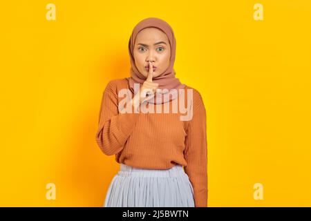 Schöne junge asiatische Frau in braunem Pullover und Hijab zeigt Stille Geste mit Finger und Blick auf Kamera isoliert über gelbem Hintergrund. Peop Stockfoto