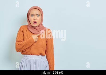 Überrascht schöne asiatische Frau in braunem Pullover und Hijab Zeigefinger im Kopierraum, zeigt Werbeprodukte und selbstbewusst Blick auf die Kamera Stockfoto