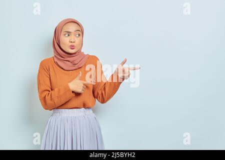 Überrascht schöne asiatische Frau in braunem Pullover und Hijab Zeigefinger im Kopierraum, zeigt Werbeprodukte und selbstbewusst Blick auf die Kamera Stockfoto