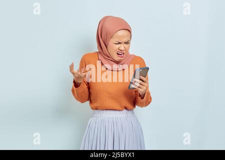 Schöne asiatische muslimische Frau in braunem Pullover und Hijab hält Handy mit wütenden Ausdruck isoliert auf weißem Hintergrund. Muslimischer Lebensstil Konz Stockfoto