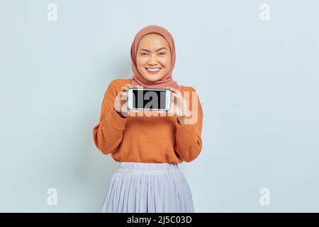 Lächelnde schöne asiatische Frau in braunem Pullover und Hijab, zeigt leeren Handy-Bildschirm isoliert auf weißem Hintergrund. Muslimische Lifestyle-Konzept Stockfoto