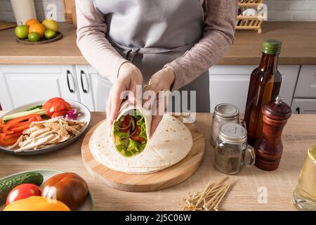Die Hände eines jungen Mädchens halten ein eingewickeltes Buritto, das zu Hause frisch gekocht wird, vor dem Hintergrund der Küche. Zutaten auf dem Tisch Stockfoto