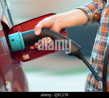 Das weibliche Modell steht neben einem Elektroauto und hält einen Ladestecker Stockfoto
