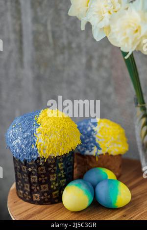 Köstliche Osterkuchen in den Farben der Flagge der Ukraine, gelb-blau gefärbte Ostereier auf einem Holztisch mit Blumen im Hintergrund. Platz f Stockfoto