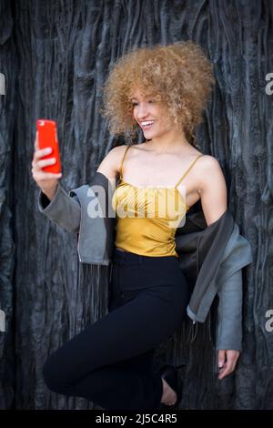 Junge lateinerin mit afro lockigen Haaren lächelt mit einem Mobiltelefon Stockfoto