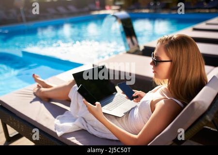 Frau, die Remote-Multitasking-Arbeit mit mehreren elektronischen Internetgeräten auf Schwimmbad Strandbett. Freiberufliche Geschäftsfrau Telearbeit mit Stockfoto