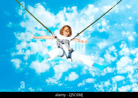 Das kleine Mädchen hüpft hoch in der Luft mit einem Bungee-Trampolin gegen den blauen Himmel mit weißen Wolken. Stockfoto