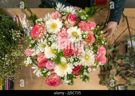 Draufsicht auf die Hände einer jungen Floristin, die eine wunderschöne Komposition aus zarten rosa Rosen, Nelken und weißen Gänseblümchen auf dem Tisch kreiert Stockfoto