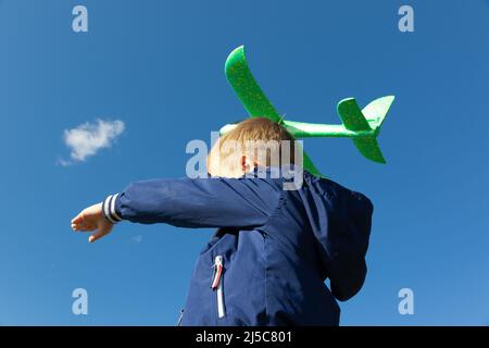 Ein sechsjähriger Vorschuljunge in blauer Jacke startet an einem Sommertag ein Spielzeug-Flugzeug in die Natur vor dem Hintergrund eines klaren blauen Himmels. Die helle Stockfoto