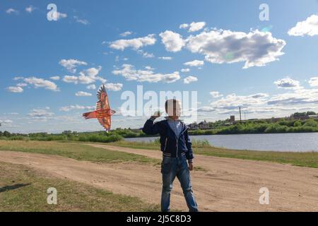 Ein sechsjähriger Vorschuljunge in blauer Jacke lässt an einem Sommertag vor dem Hintergrund eines klaren blauen Himmels einen Drachenvögel in die Natur fliegen. Die helle Stockfoto