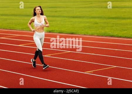 Eine wunderschöne Sportlerin, die im Stadion läuft, trainiert, sich in Form hält, Sportbekleidung trägt, ein weißes Oberteil und eine weiße Hose trägt, empfiehlt, Sport zu treiben Stockfoto