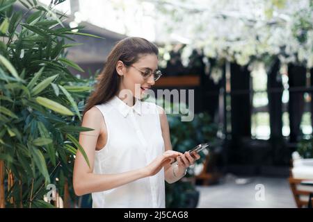 Junge Frau mit einem Smartphone, die in der Halle eines gemütlichen Restaurants steht. Stockfoto