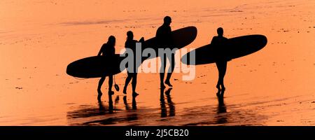 Ein Panoramabild einer Gruppe von Surfern, die ihre Surfbretter tragen und am Fistral Beach entlang spazieren, der von einem intensiven Sonnenuntergang in Newquay in Cornw umragt wird Stockfoto