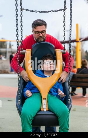 Ein entspanntes Kind mit einer Behinderung spielt auf angepasster Schaukel mit seinem Vater auf einem Spielplatz Stockfoto