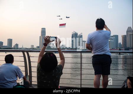 07/21/2018, Singapur, Republik Singapur, Asien - Zuschauer beobachten, wie Hubschrauber der Luftwaffe (RSAF) während eines Trainingsfluges eine riesige Staatsflagge über Wolkenkratzer in Marina Bay fliegen, um sich auf den bevorstehenden Nationalfeiertag am 9. August vorzubereiten. [Automatisierte Übersetzung] Stockfoto