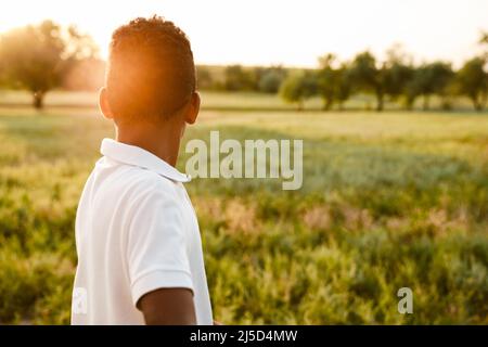 Schwarzer Junge in weißem Hemd, der den Sonnenuntergang anschaut, während er auf dem Sommerfeld steht Stockfoto