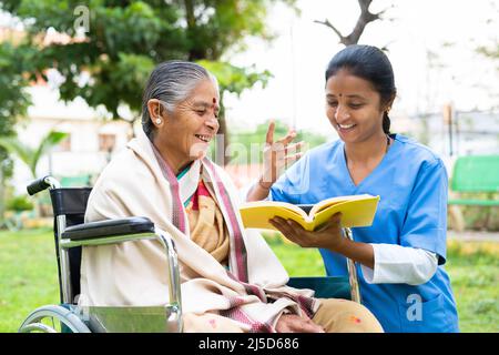 Krankenschwester und ältere Frau auf dem Rollstuhl lachen beim Lesen von Buch oder Roman im Park - Konzept der Entspannung, Beruf, Therapie oder Behandlung Stockfoto